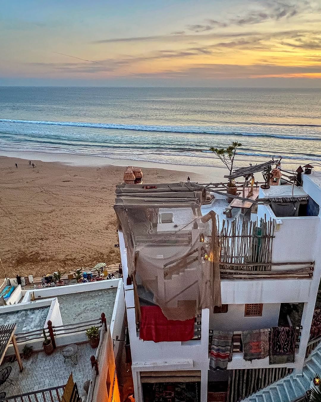 Golden-hour sunset over Taghazout beach from a hand-built rooftop terrace with lanterns and woven shade, Morocco