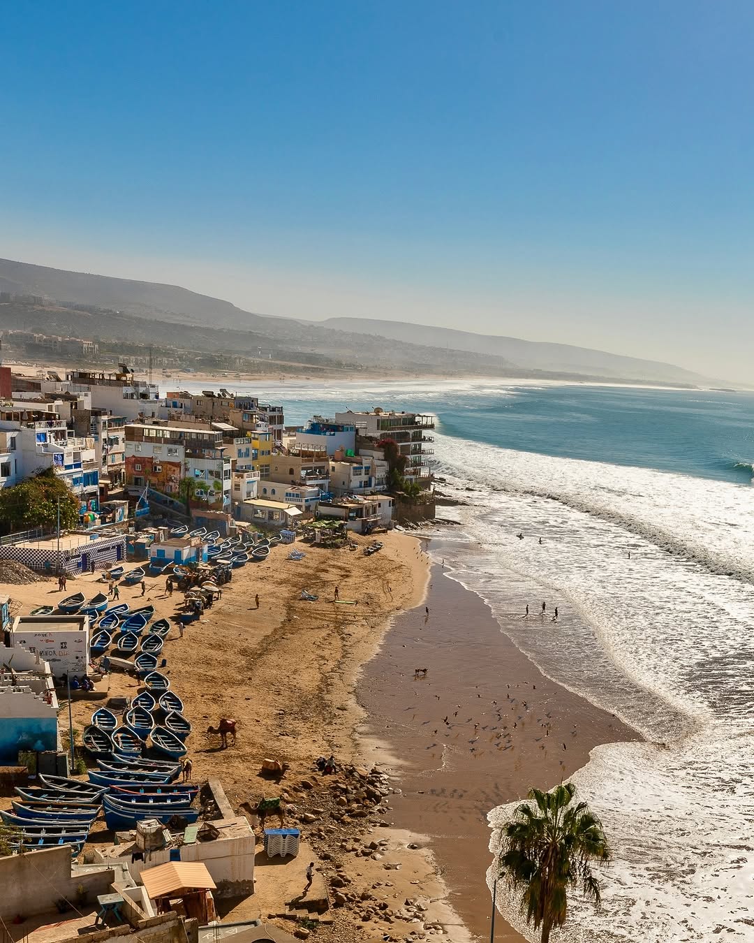 Hand-painted blue fishing boats lined on Taghazout beach with surfers in the Atlantic waves behind, Morocco
