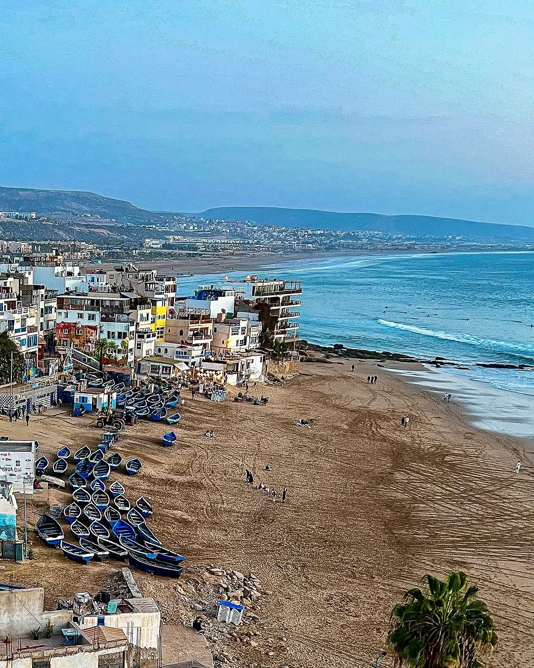 Aerial view of the Taghazout coastline with blue fishing boats, the white village and Anti-Atlas mountains, Morocco