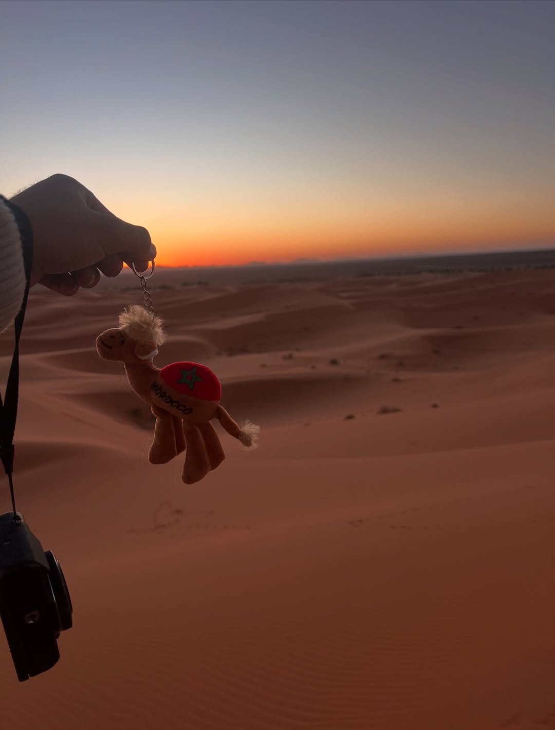 Sahara Souvenir Morocco camel keychain held in front of Merzouga sunset dunes