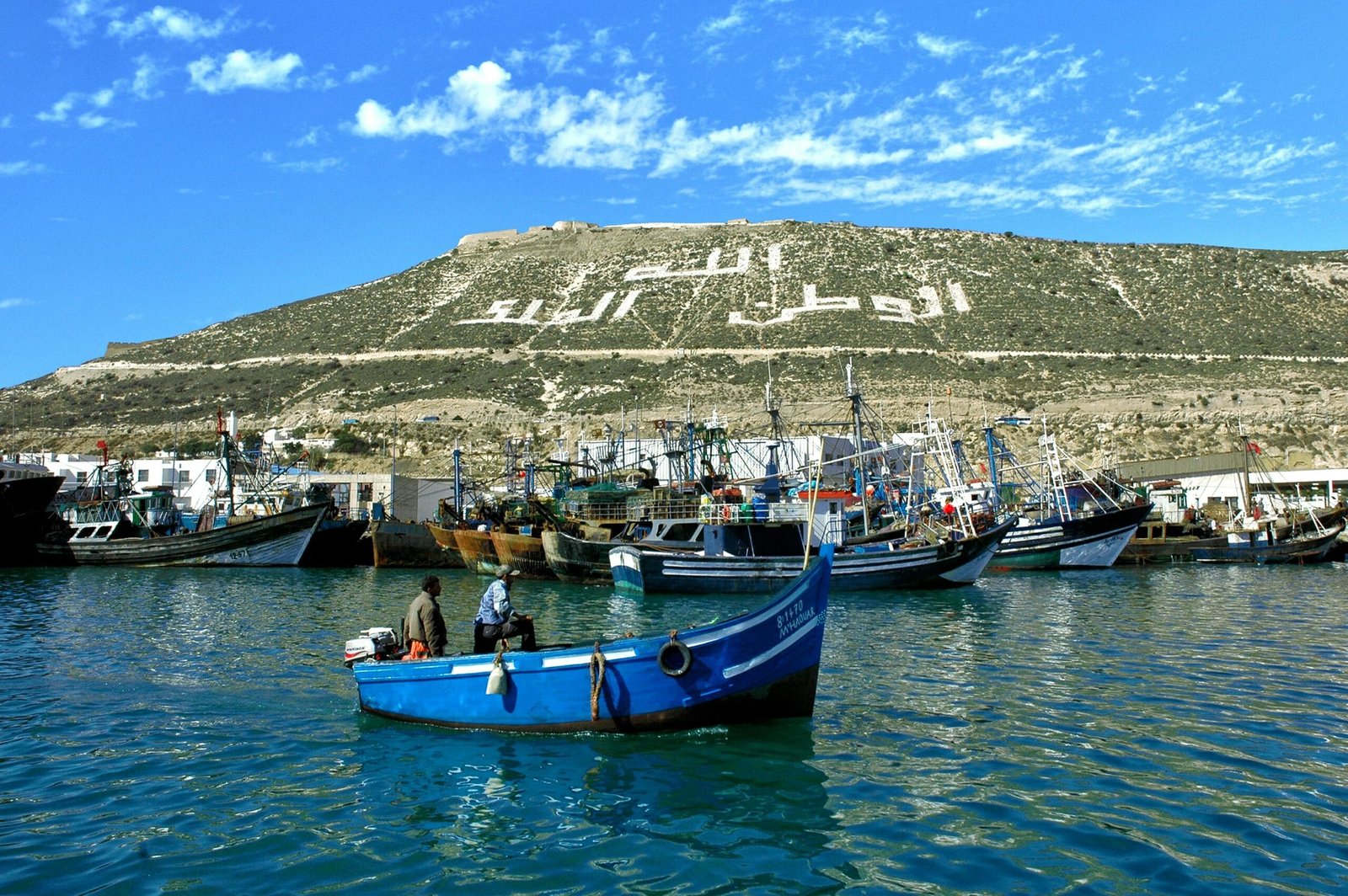 Fishing boats anchored in Agadir port under a hill with a prominent Arabic inscription.