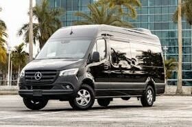 Black Mercedes-Benz Sprinter van in urban setting with modern building and palm trees.